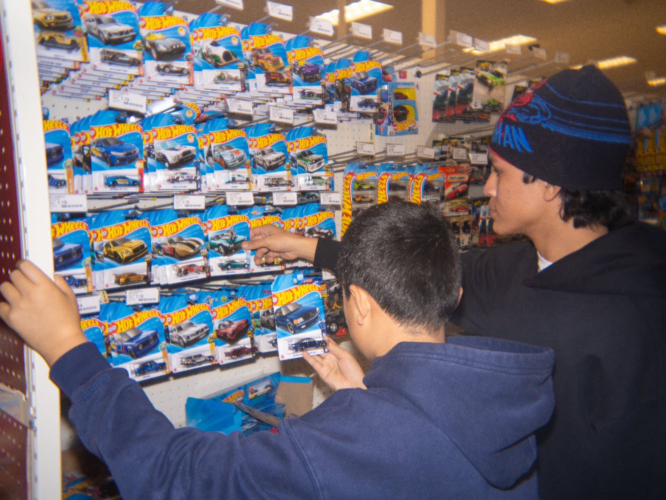 Two boys browsing Hot Wheels die-cast toy cars on a store display shelf, featured for 3G Unique Decor's free Hot Wheels car promotion on orders over $100.