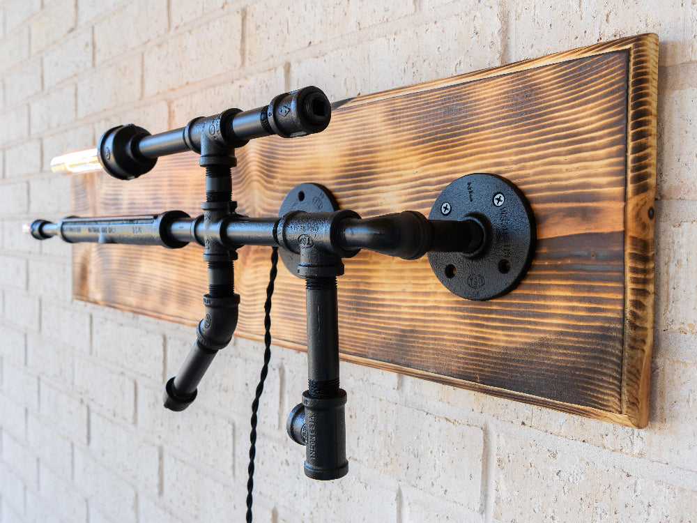 Wall-mounted light fixture with black pipes and wooden panel on a brick wall.