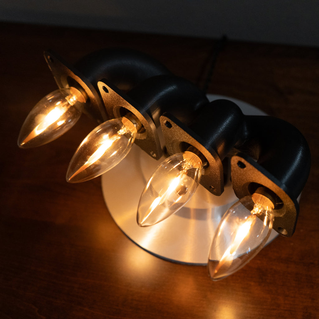 Handcrafted automotive desk lamp made from a black 4-cylinder exhaust manifold with four warm lit candle flame bulbs on a metal base.