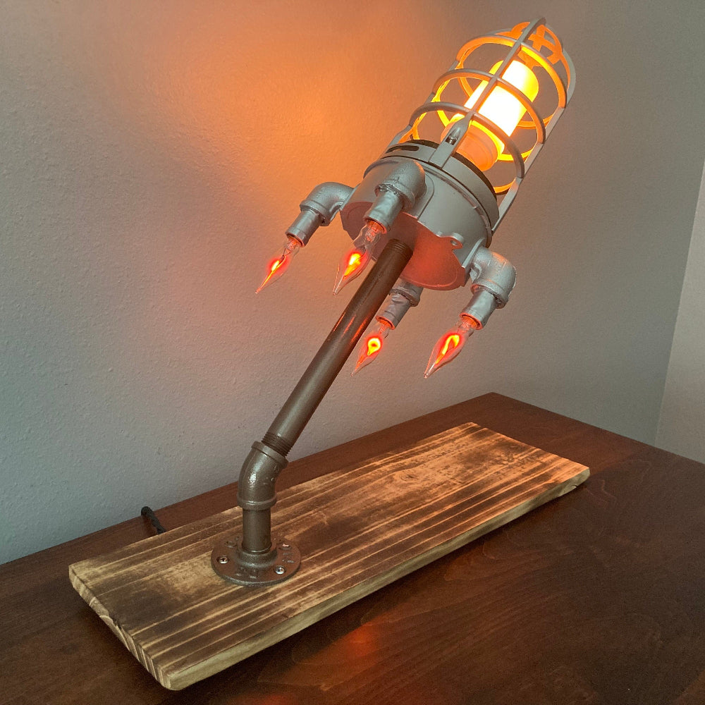 Rocket lamp mounted on burnt wood base with galvanized pipe support for stable display.